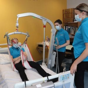 two female students observe mock patient sitting on bed via a pulley