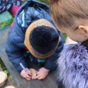 child decorates wood (story slice), watched by female student