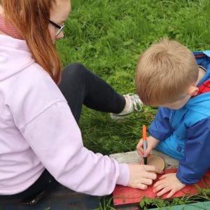 child decorates wood (story slice), watched by female student