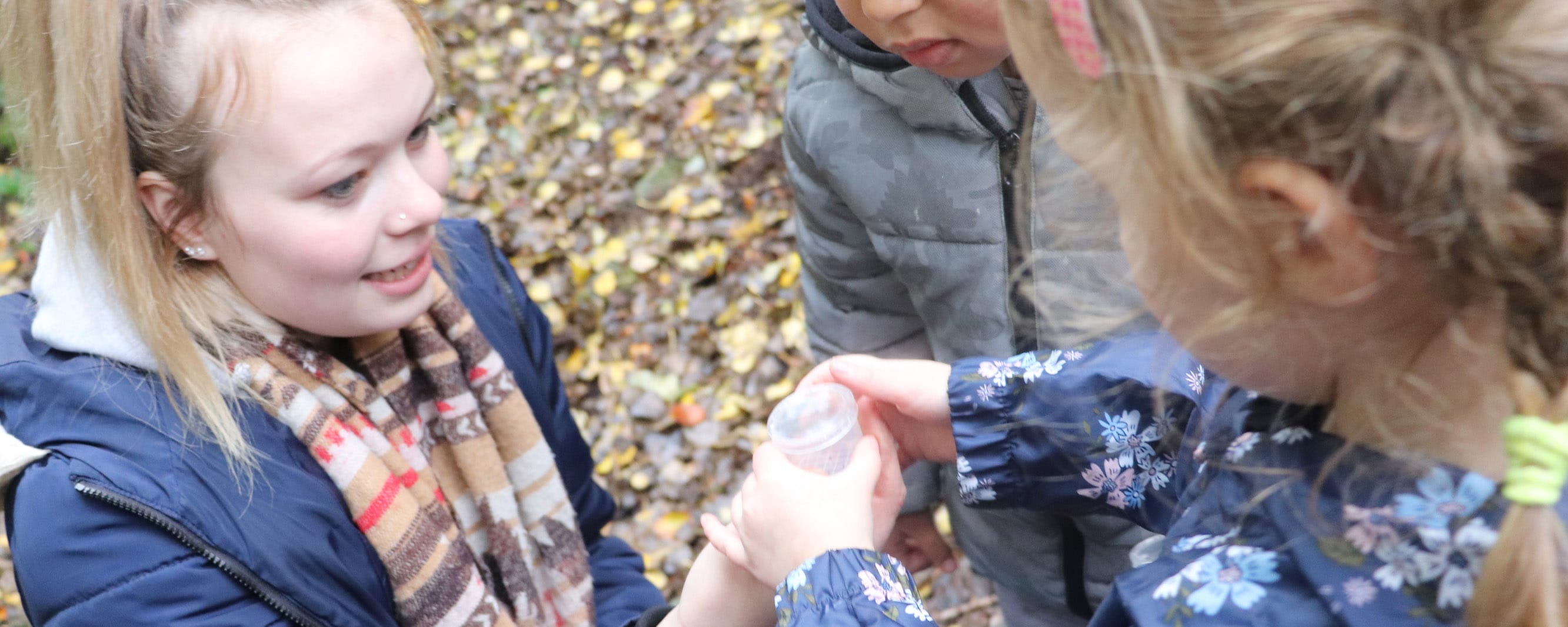 female student hands bug hunting container to girl, watched by a boy