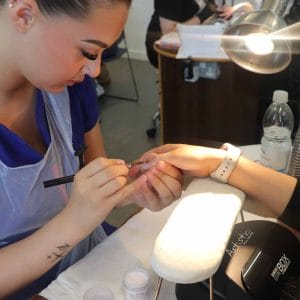 female student nail technician applies brush to nail of a client with lights in background