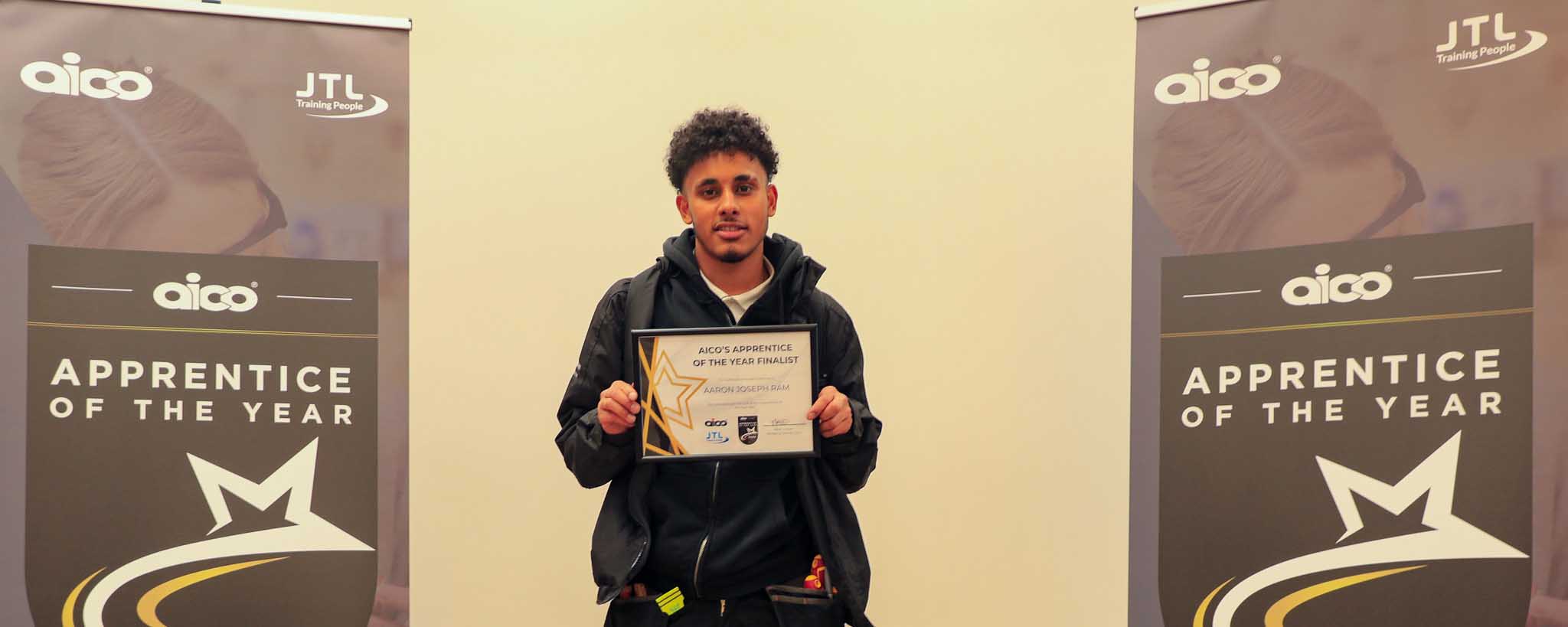 male apprentice poses with certificate award by banner stands facing