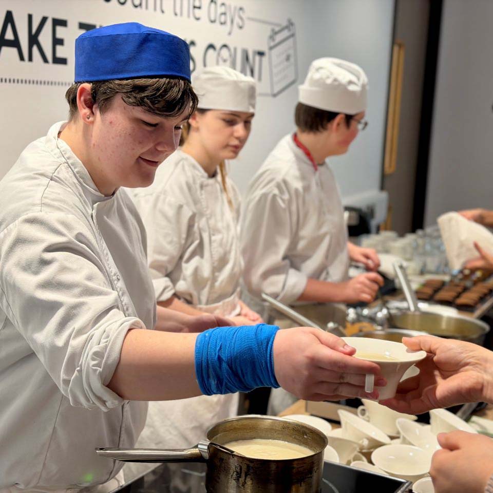 male student hands soup bowl to diner with two other students at work in the background