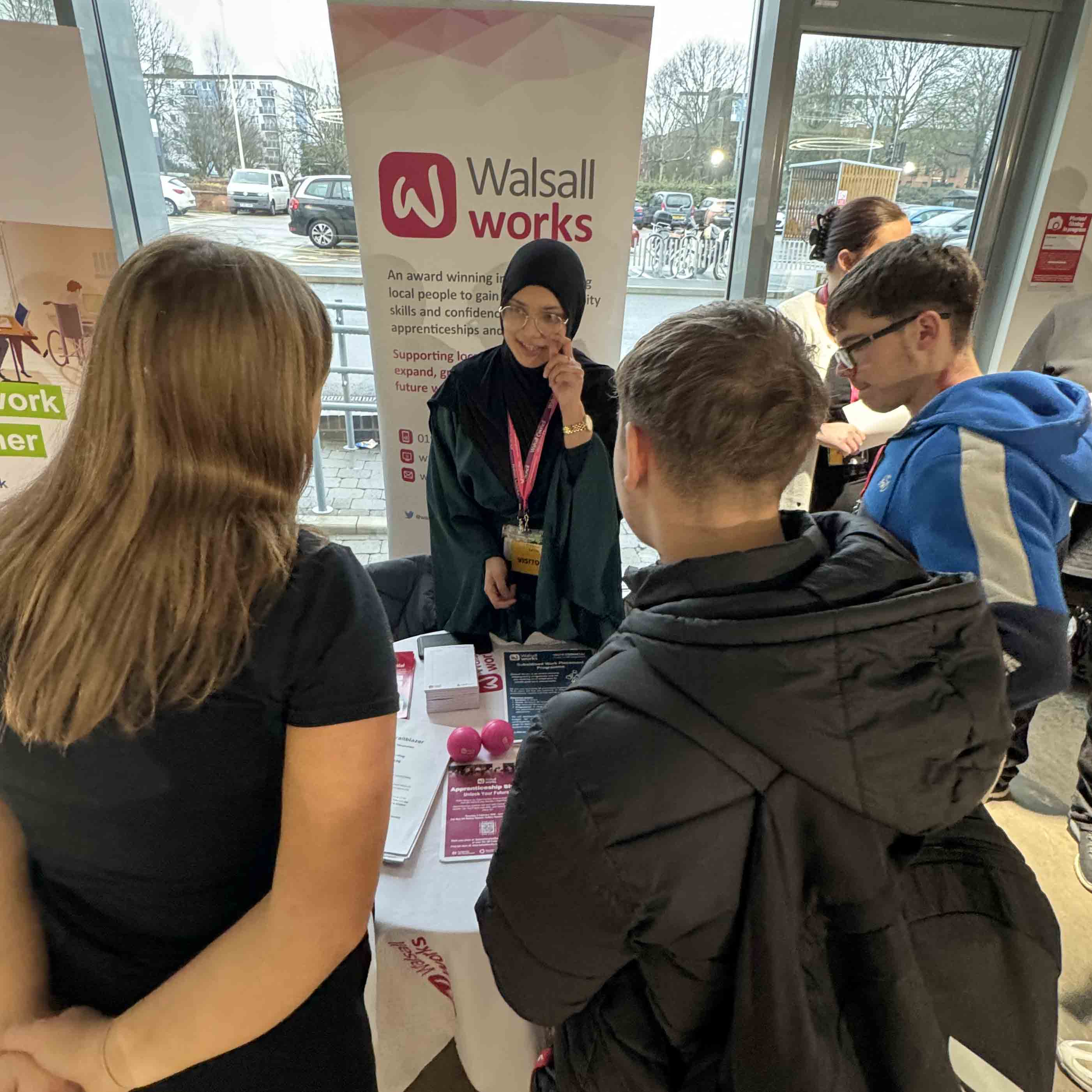 female Walsall Works representative at exhibition stand speaks to three students 